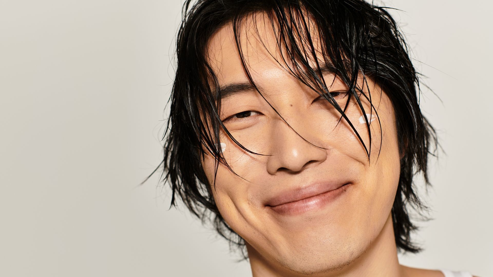 特寫鏡頭，一位微笑的黑髮男子臉上塗著兩滴潤膚露。