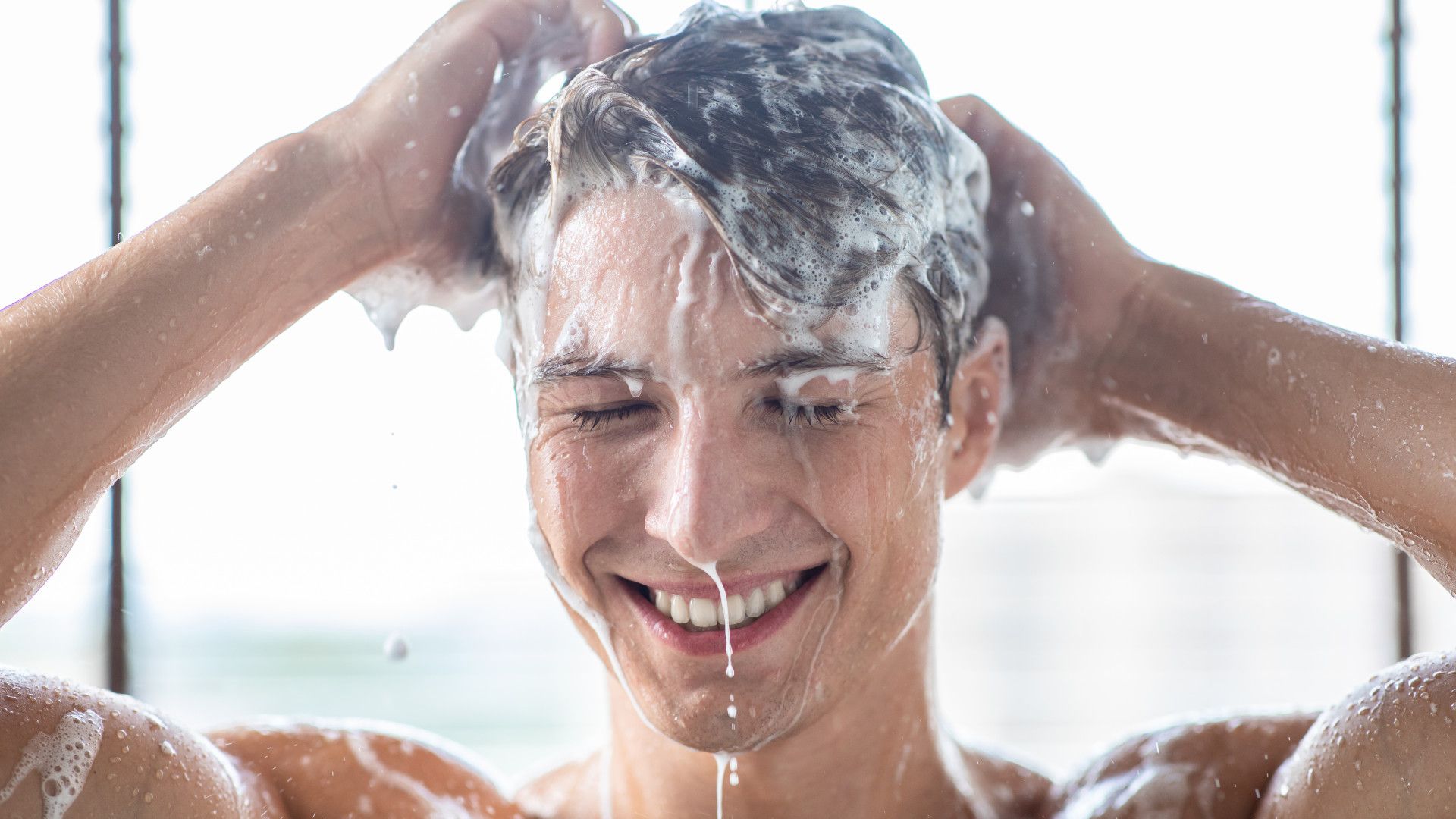 一個微笑的人在淋浴間用洗髮精洗頭，閉著眼睛，水和泡沫順著臉頰流淌下來。